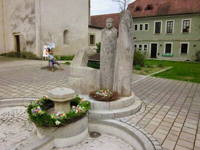 Während Stups auf dem Platz für Aufregung sorgt, gibt es eine Sache, die von ihm verschont bleibt: der festlich geschmückte Brunnen. Ein liebevoll gebundener Osterkranz und handbemalte Eier, die von den Damen des Frauenstammtisches gestaltet wurden, zieren das Herzstück des Kirchplatzes. Manche Dinge sollten einfach bleiben, wie sie sind.